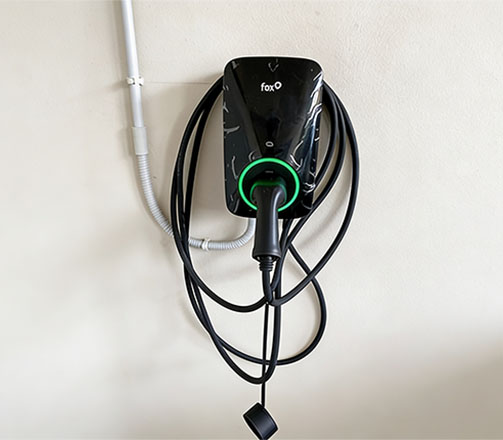 Wallbox nera a marchio Fox per la ricarica di veicoli elettrici, installata a parete, con anello luminoso a LED verde acceso e cavo di ricarica nero avvolto intorno alla struttura.