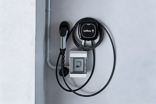 Stazione di ricarica nera a marchio Wallbox installata a parete, affiancata da un piccolo quadro elettrico di protezione bianco e dal supporto a muro su cui è agganciata la presa del cavo di ricarica.