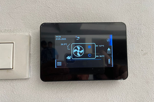 Display digitale nero touch screen installato a parete, con schermo acceso che mostra grafiche e icone blu relative al funzionamento di una pompa di calore, affiancato a una placca per interruttori bianca.