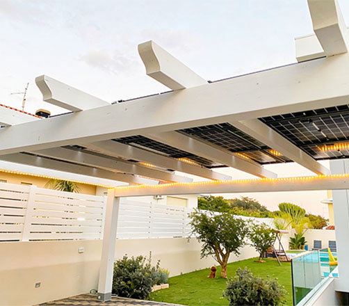 Struttura di una pergola in legno bianco con pannelli solari fotovoltaici integrati come copertura, situata nel giardino di una casa.