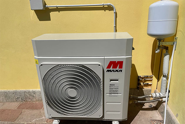 Unità esterna bianca di una pompa di calore a marchio MAXA installata su una pavimentazione esterna a ridosso di un muro giallo, con tubazioni isolate e un piccolo vaso di espansione bianco sulla destra.